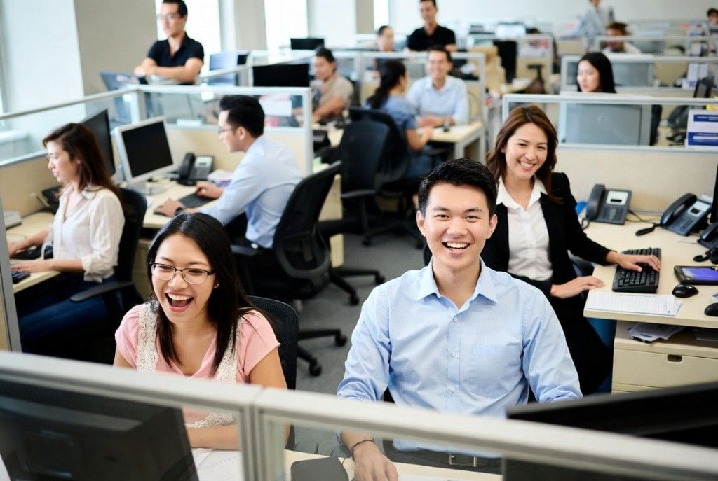 A smiling back-office teammate at his workstation in a busy open-office with coworkers also working behind him — the kind of dedicated back-office team Online VA Team places with growing US businesses