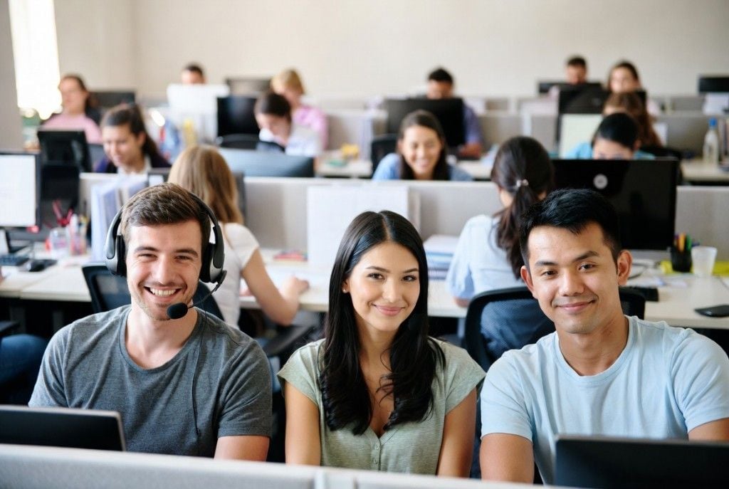 Three smiling teammates at the front of a busy bullpen — a man in a headset, a woman in a green tee, and a man in a light blue tee — with coworkers working at desks behind them. The kind of real team that'll read your message and reply.