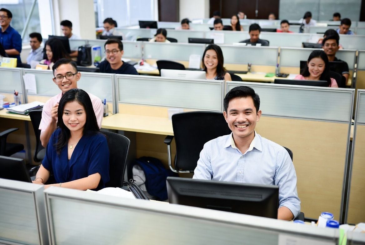 A bright open-floor office where three smiling teammates in the foreground are taking a moment from work — a man in a light blue button-down at center, two women on either side — with a busy bullpen of teammates working in cubicles behind them. The kind of real team Online VA Team places with growing US businesses.