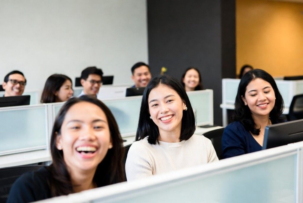 A team of smiling Filipino teammates working at their cubicles in a busy modern office, with a woman laughing in the foreground — the kind of dedicated remote teammates Online VA Team places with growing US businesses