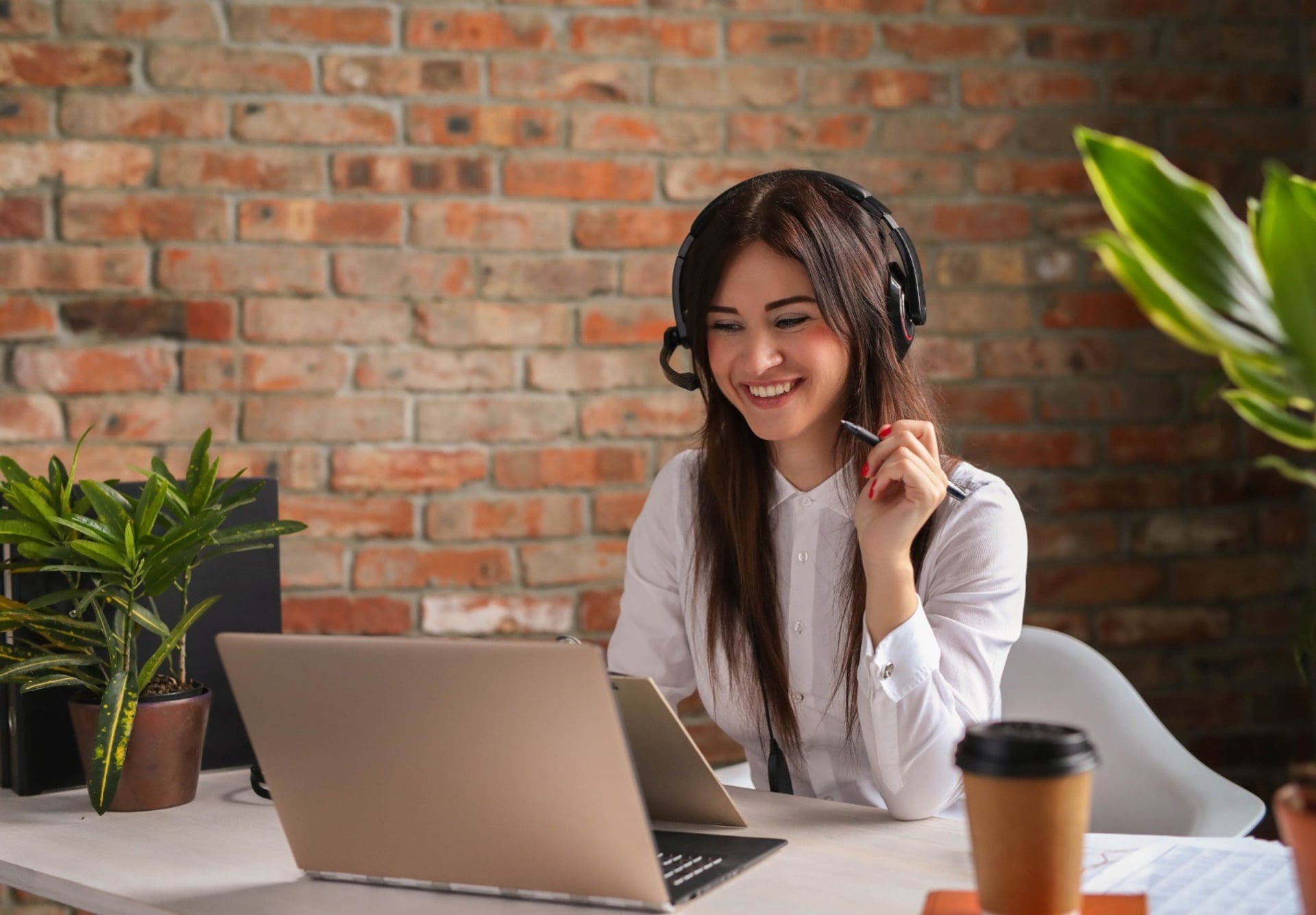 A smiling customer service teammate in a headset at her laptop, mid-call, in a bright brick-walled office