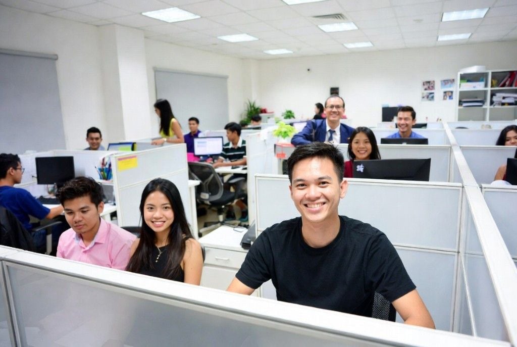 A team of three smiling teammates at their desks with a friendly manager standing behind in a modern office