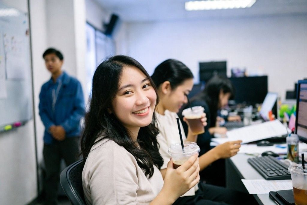 Smiling bookkeeper in a cream top holding an iced coffee at her desk, teammates working alongside her in a sunlit open office