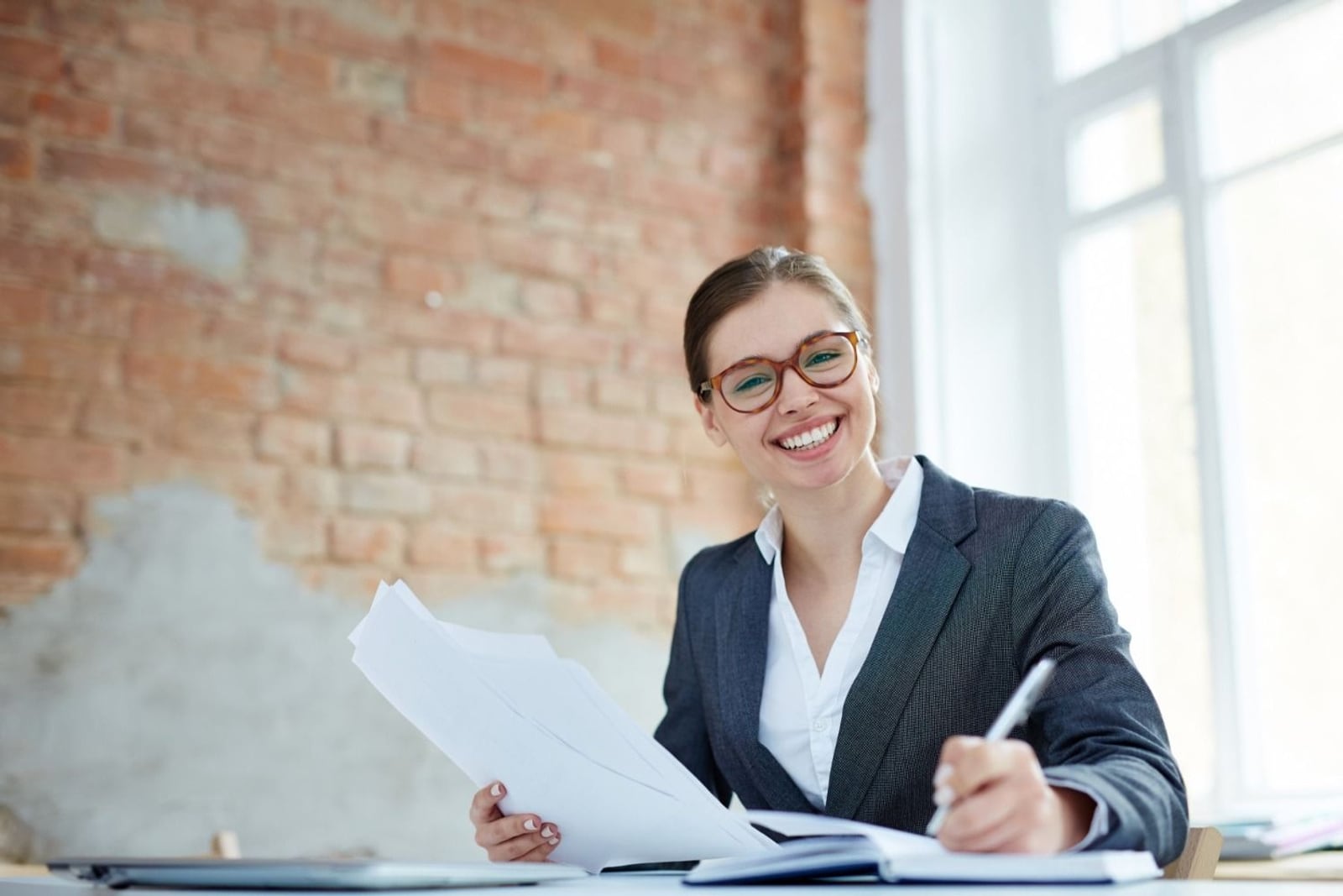 A smiling teammate at her desk holding a printed statement and a pen, mid-way through a set of books, in a bright brick-walled office